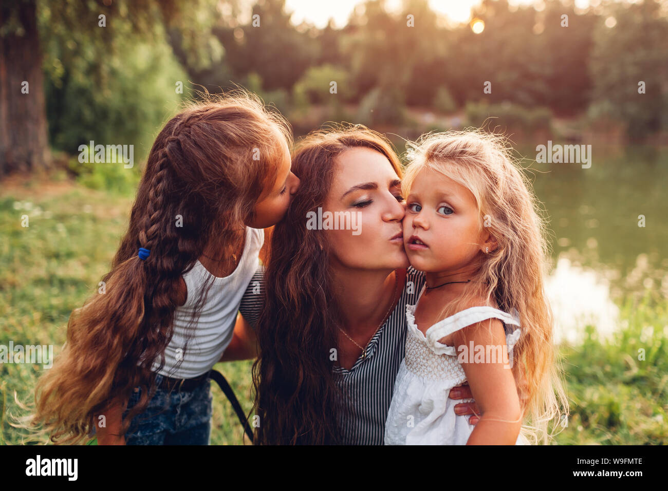 Mother and her daughters hugging and kissing outdoors. Family walking by summer river at sunset. International Childrens Day Stock Photo