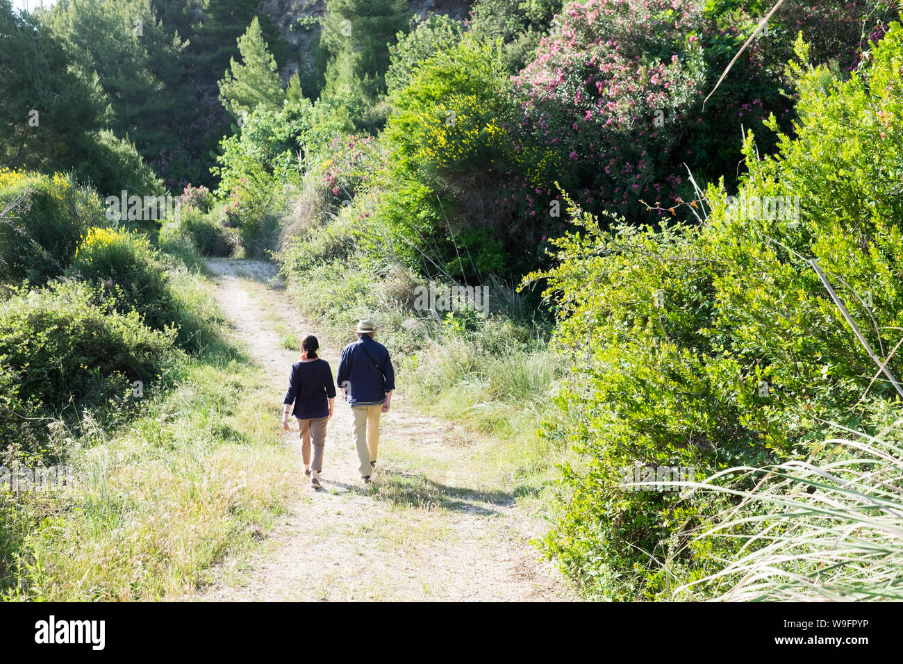 An adventurous couple in their late 60's early 70's walk down a path lined with wildflowers and shrubs in Kefalonia Greece while on holiday. Stock Photo