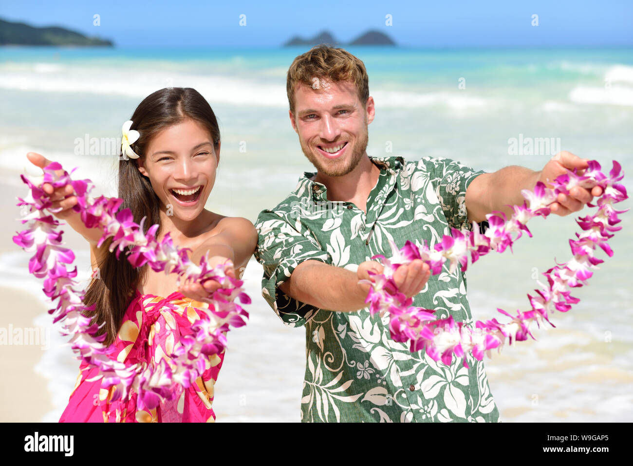 Welcome to Hawaii - Hawaiian people showing giving leis flower necklaces as welcoming gesture for tourism. Travel holidays concept. Asian woman and Caucasian man on white sand beach in Aloha clothing. Stock Photo