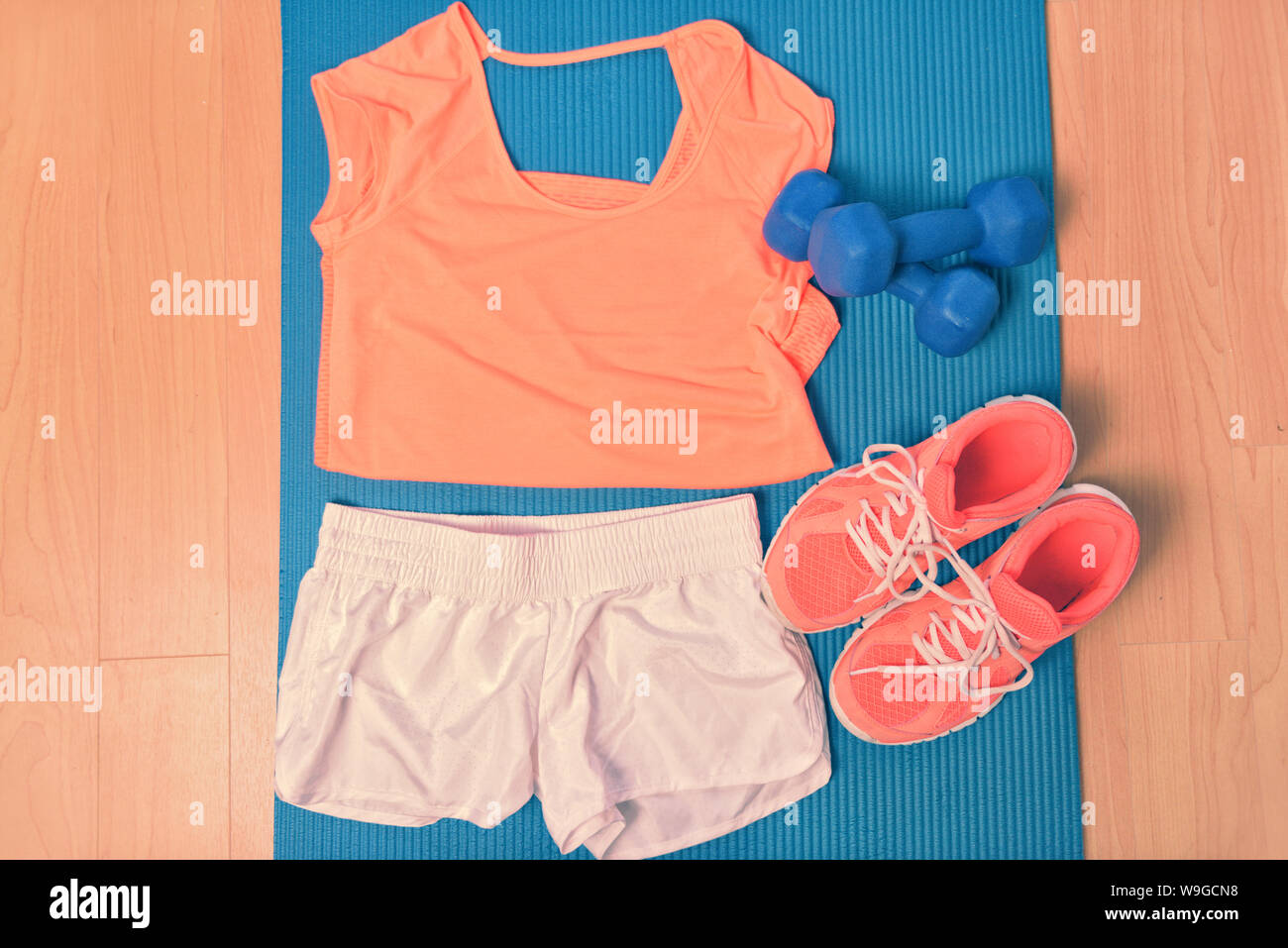 Workout clothes - fitness outfit and running shoes. Overhead of clothing ready for lifting weights at the gym or at home, laying on a yoga mat on the floor. Matching orange t-shirt and sneakers. Stock Photo