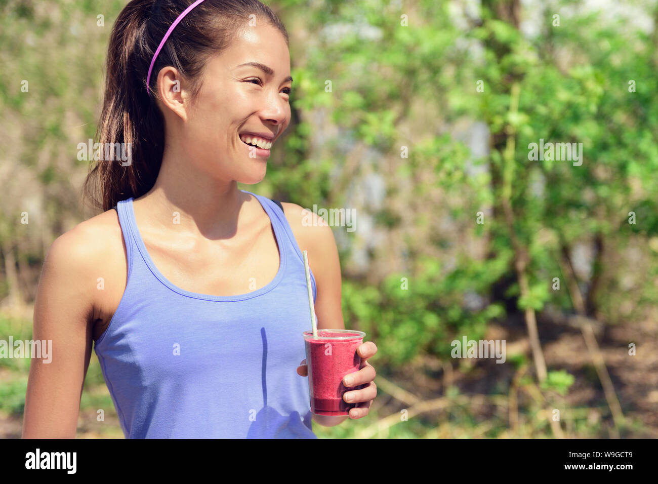 Healthy Asian woman drinking fruit smoothie drink in outdoor forest park during summer. Young fit girl holding plastic cup clean eating for detox cleansing with berry or beet juice as part of a diet. Stock Photo