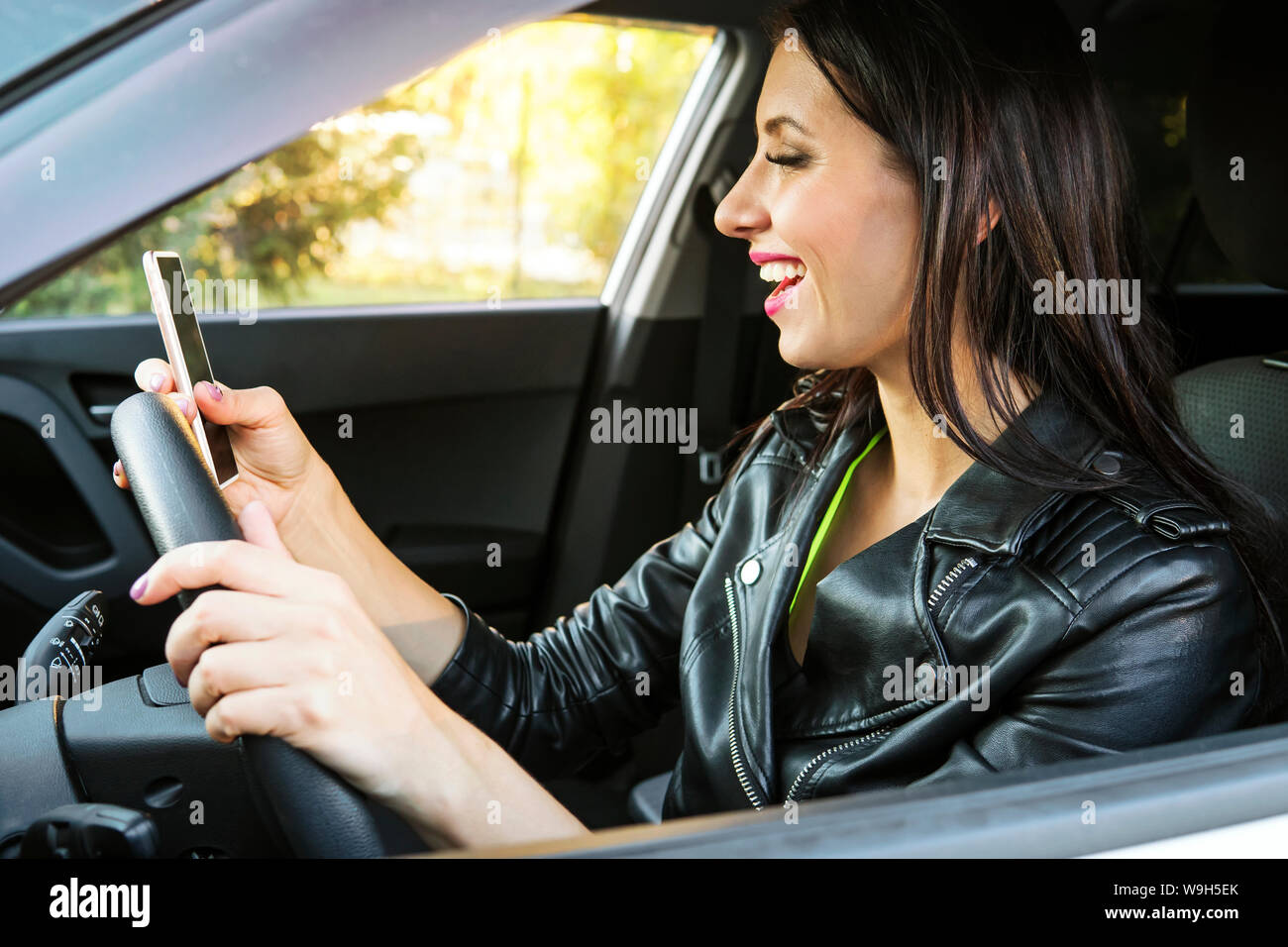 Happy smiling woman holding her smart phone sitting inside a car. Female driver laughing reading message and chatting in mobile. Girl looks at her Stock Photo