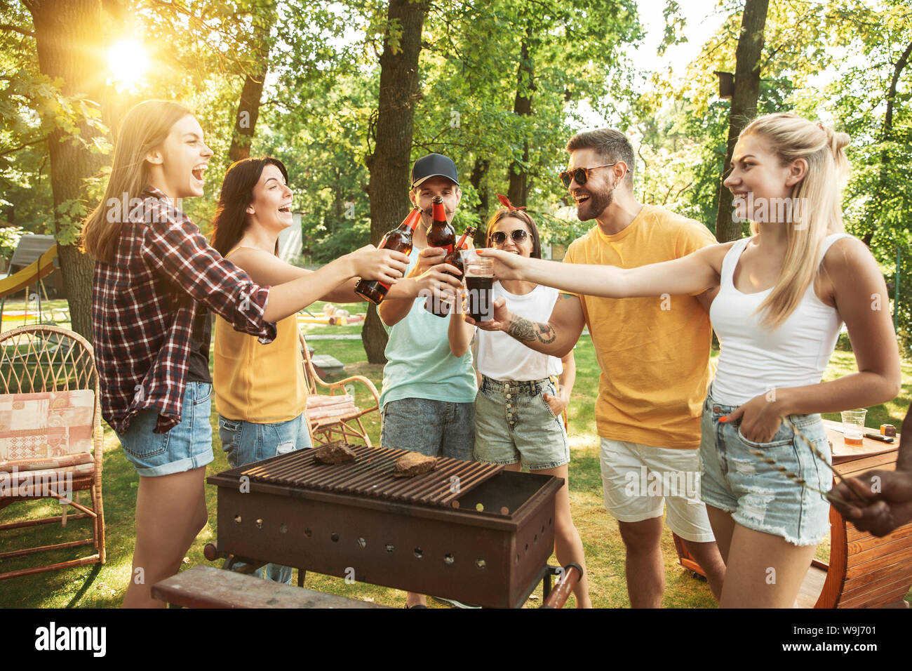 Group of happy friends having beer and barbecue party at sunny day. Resting  together outdoor in a forest glade or backyard. Celebrating and relaxing,  laughting. Summer lifestyle, friendship concept Stock Photo -