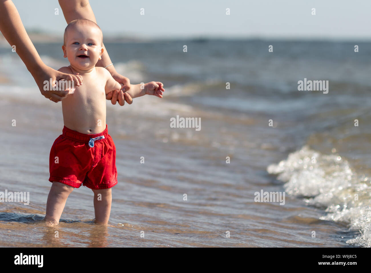 Funny baby boy for the first time by the sea trying to walk in the water with the help of her mom Stock Photo
