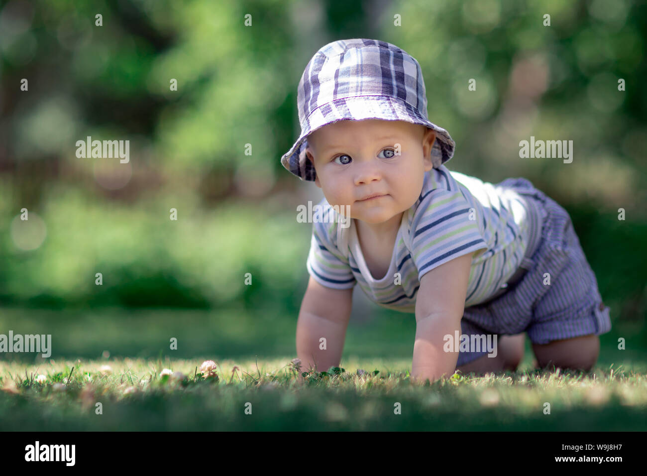 Baby with nice blue cap crawling on the green grass in the garden and smiles Stock Photo