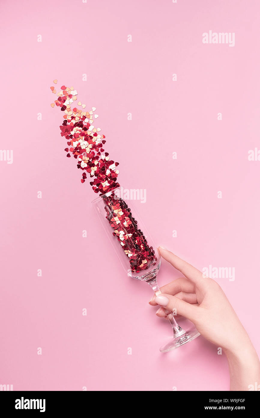 Female hand holding champagne glass with vibrant red heart-shaped confetti poured out of it on pink background. Flat lay. Love and celebration concept Stock Photo