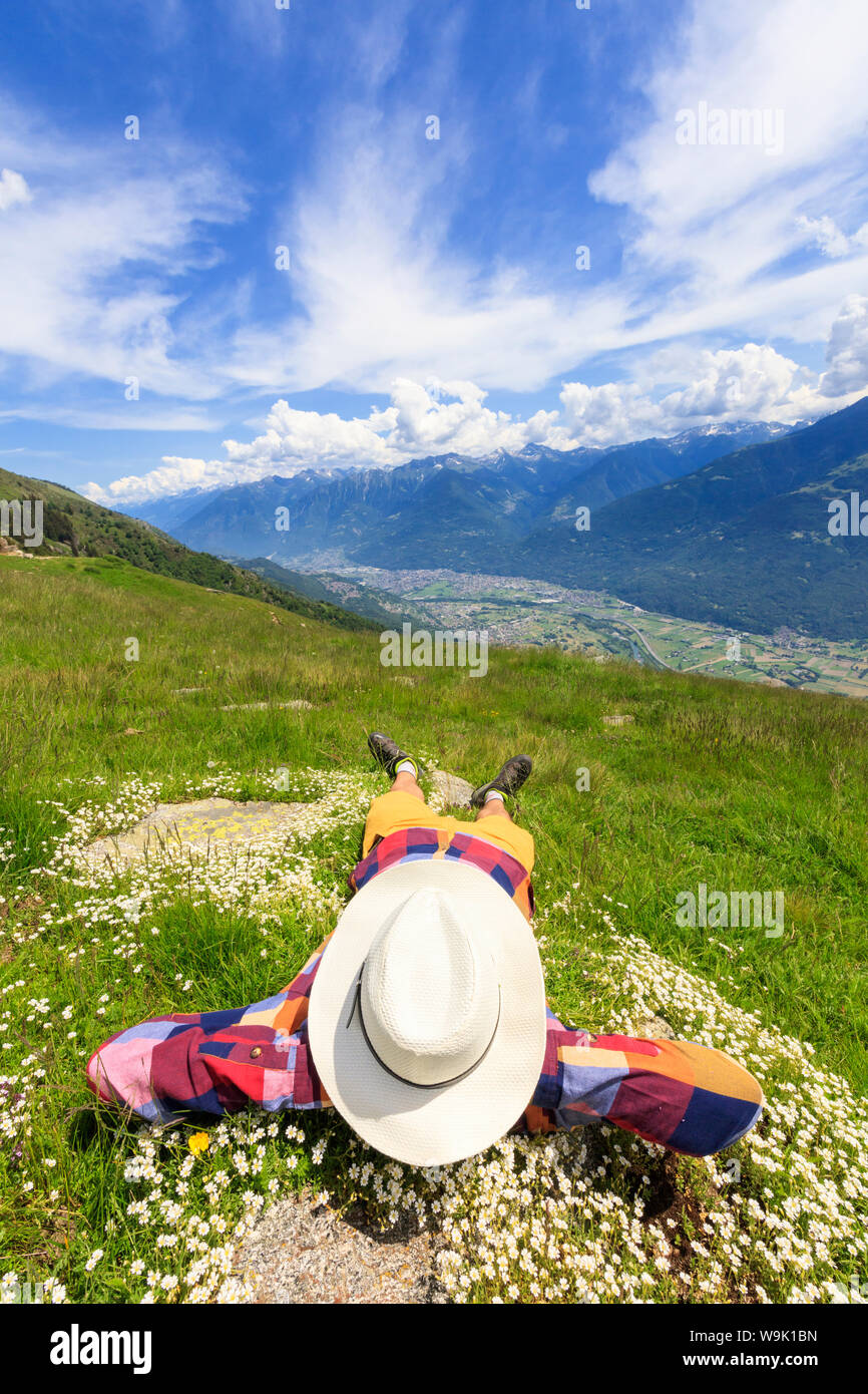 Rear view of man with hat lying on grass, Alpe Bassetta, Valtellina, Sondrio, Lombardy, Italy, Europe Stock Photo