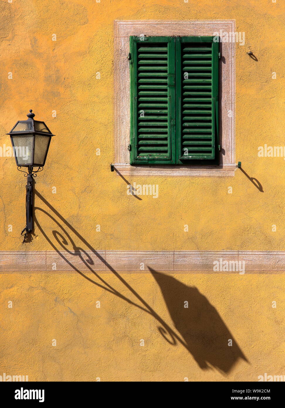 Architectural detail showing green wooden shutter on yellow wall with ornate wall lamp and shadow, San Quirico d'Orcia, Tuscany, Italy, Europe Stock Photo