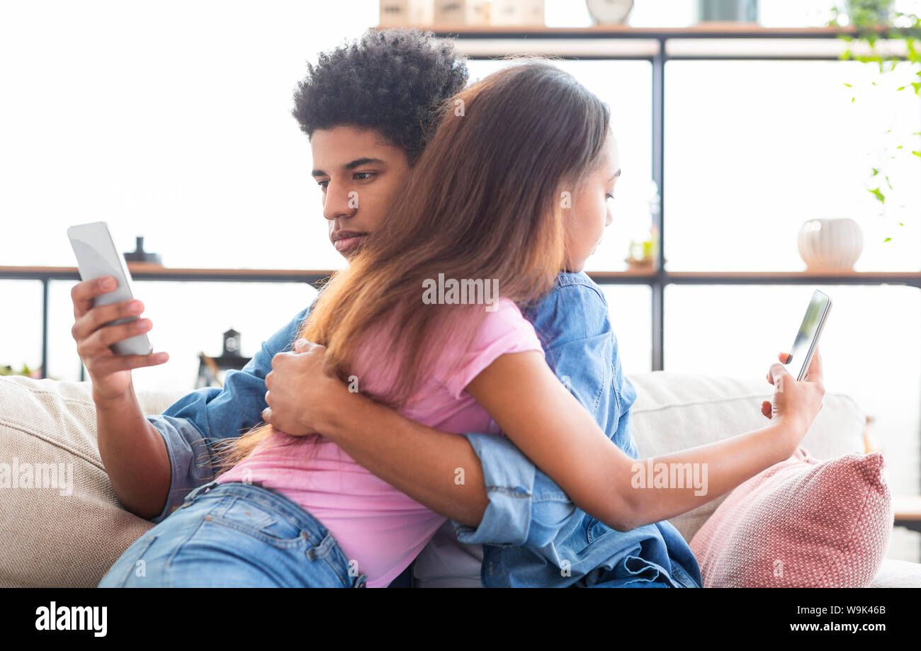 Teen couple cuddling but looking at mobile phones over shoulder. Stock Photo