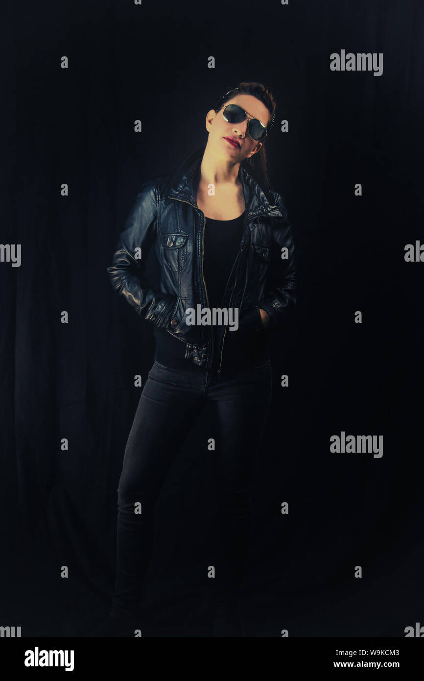 A woman in a black leather jacket and sunglasses standing and looking cool. Stock Photo
