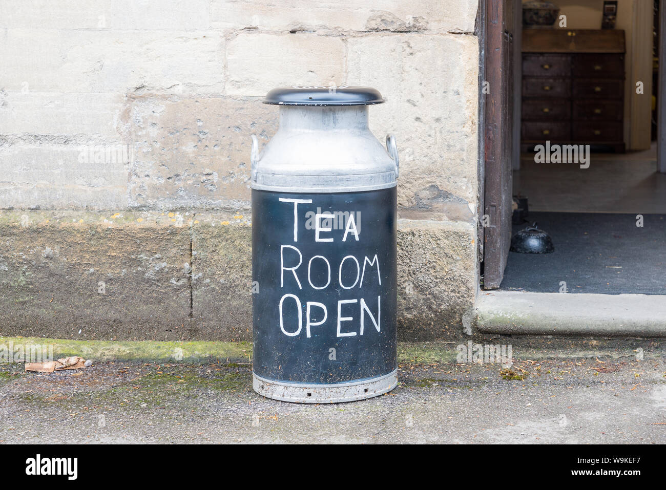 Traditional aluminium milk churn outside cafe used as sign saying 'Tea Room Open' in Abbey Terrace, Winchcombe,Gloucestershire,UK in the Cotswolds Stock Photo
