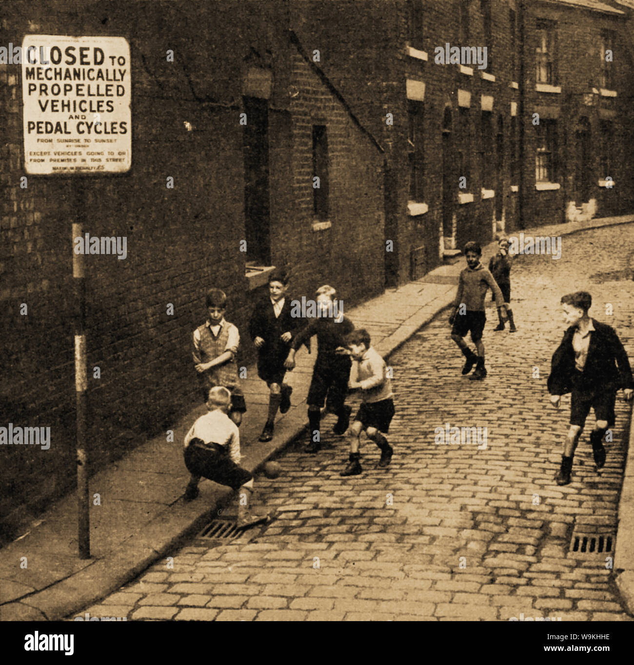 Circa 1940's - British school children from the north of England play football in the street - A sign prohibits all vehicles except those of residents to enter the cobbled street Stock Photo