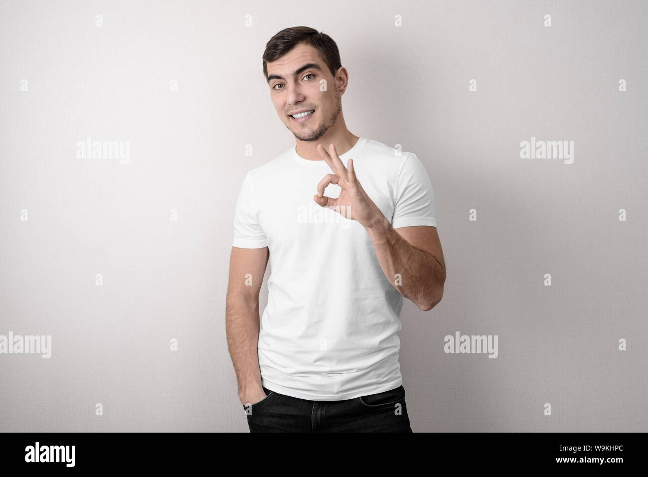 Young European man in white t-shirt on grey background shows OK sign, studio shots Stock Photo