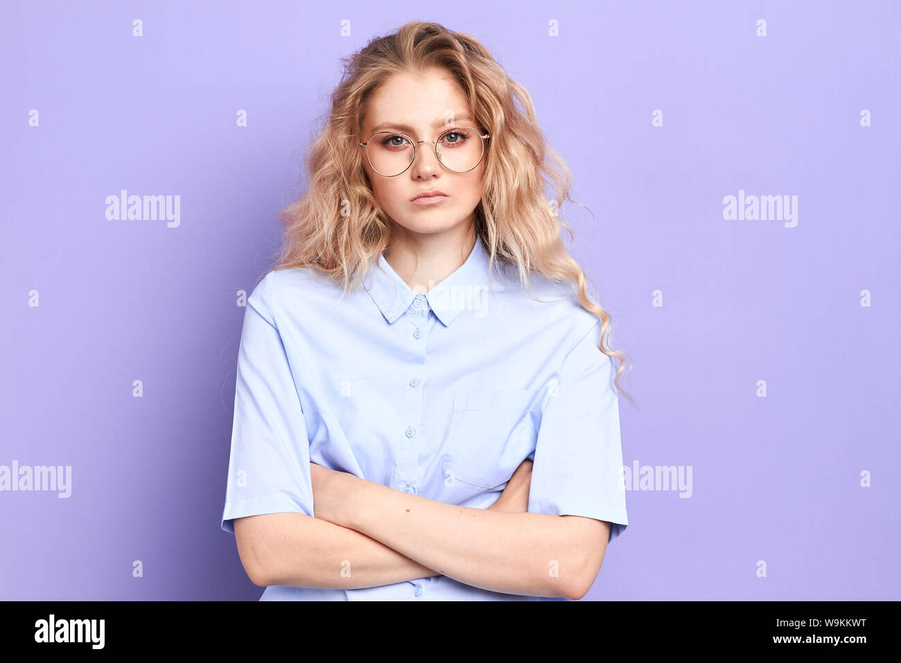 Beautiful confident serious young woman in eyewear looking at camera and keeping arms crossed while standing against blue background, strict teacher Stock Photo