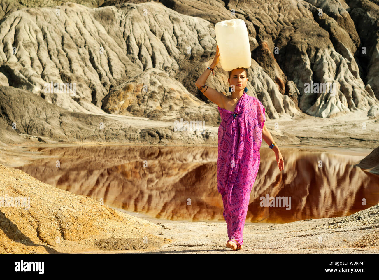 girl of oriental appearance in a sari carries on her head a large plastic canister  with water over a desert area Stock Photo