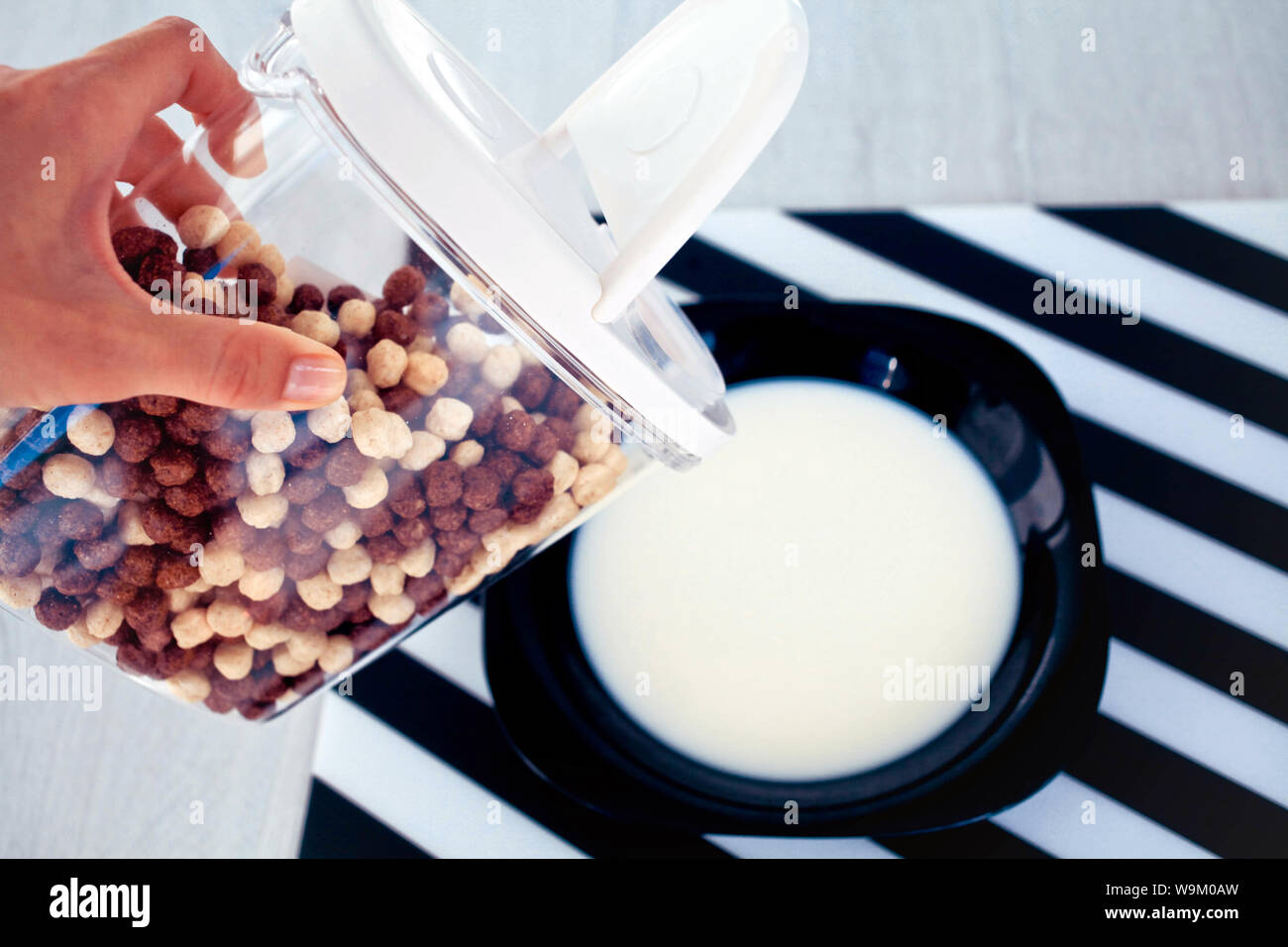 female hand holds in hand the container chocolate cereal and corn balls and a black plate with milk. Stock Photo