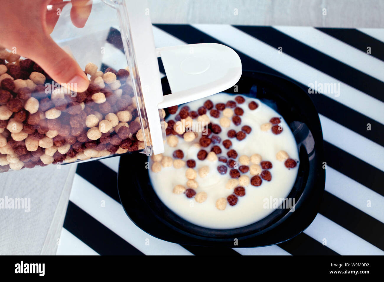 female hand fills from the container chocolate cereal and corn balls in a black plate with milk. Stock Photo