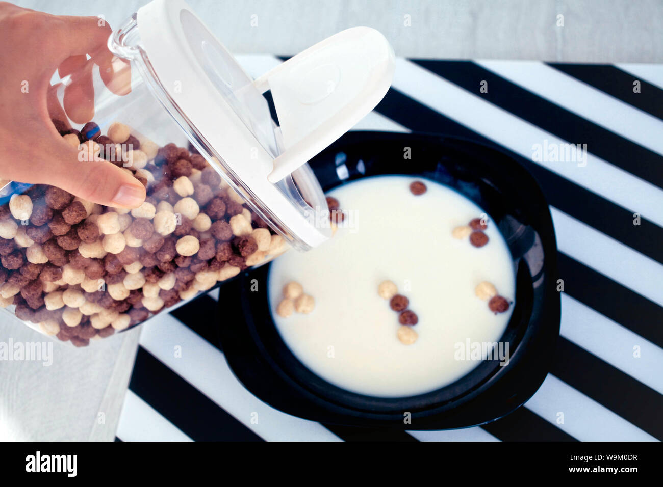 female hand fills from the container chocolate cereal and corn balls in a black plate with milk. Stock Photo