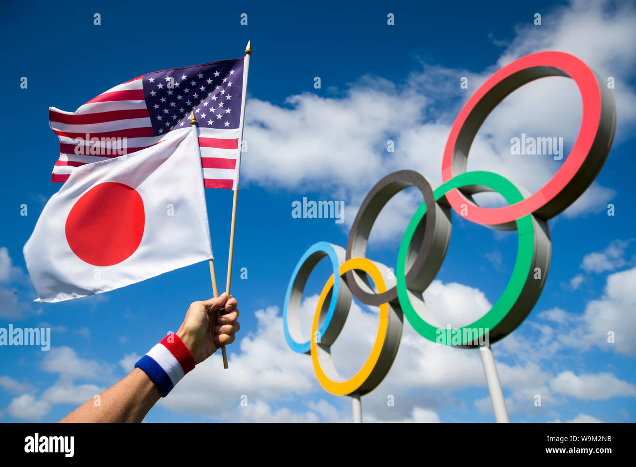 LONDON - APRIL 19, 2019: Hand wearing red white and blue wristband holds Japanese and American flags together in front of Olympic Rings. Stock Photo