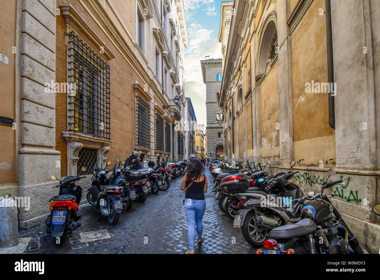 A woman walks alone down a narrow side street with many motorcycles parked on either side in the historic center of Rome, Italy Stock Photo