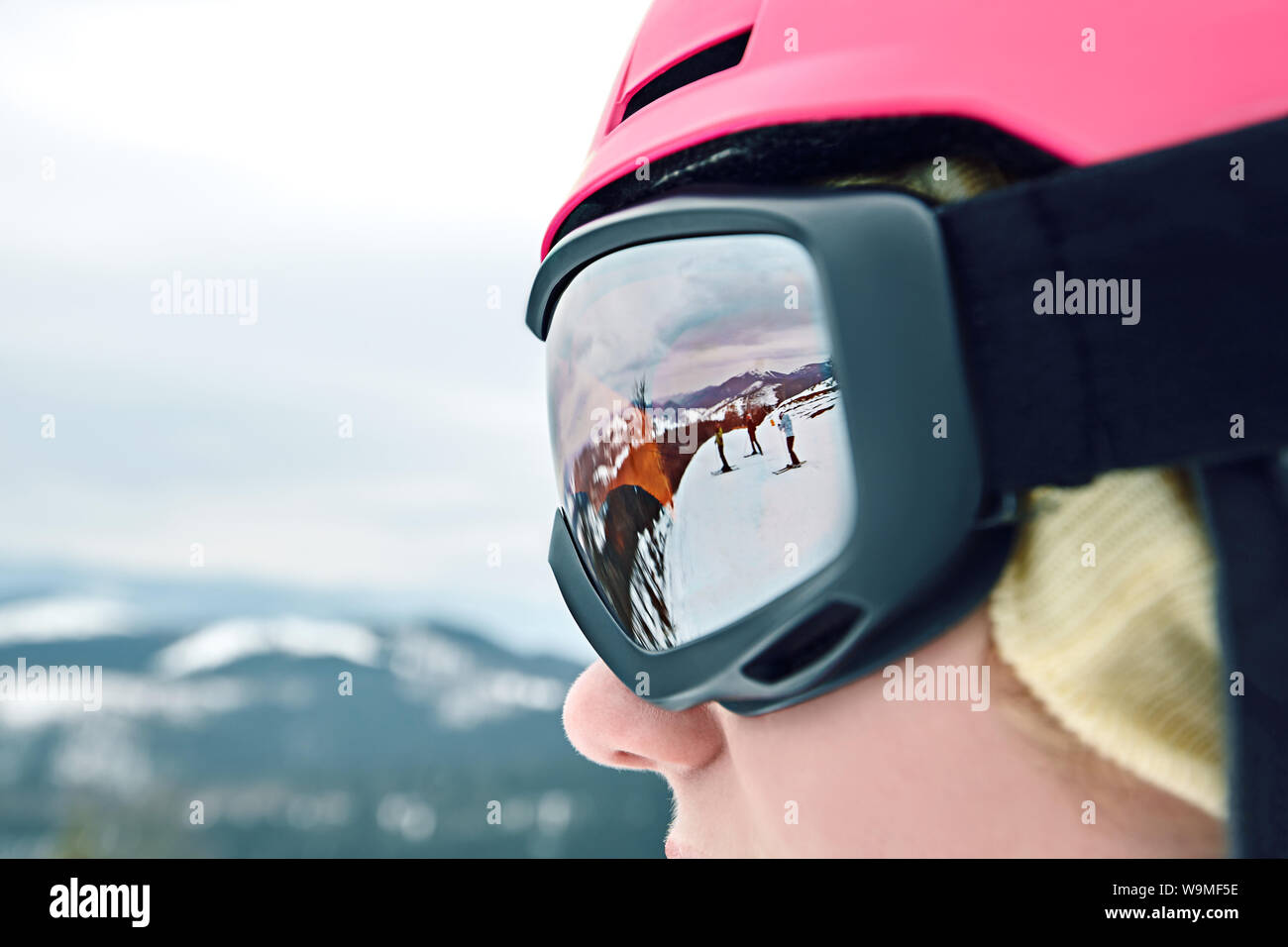 Close up portrait of snowboarder woman at ski resort wearing helmet and goggles with reflection of mountains. Stock Photo