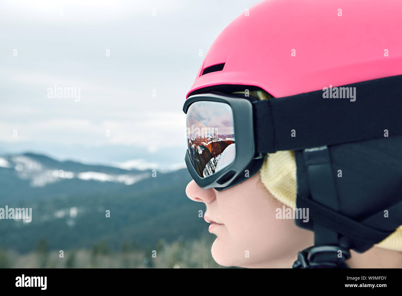 Close up portrait of snowboarder woman at ski resort wearing helmet and goggles with reflection of mountains. Stock Photo