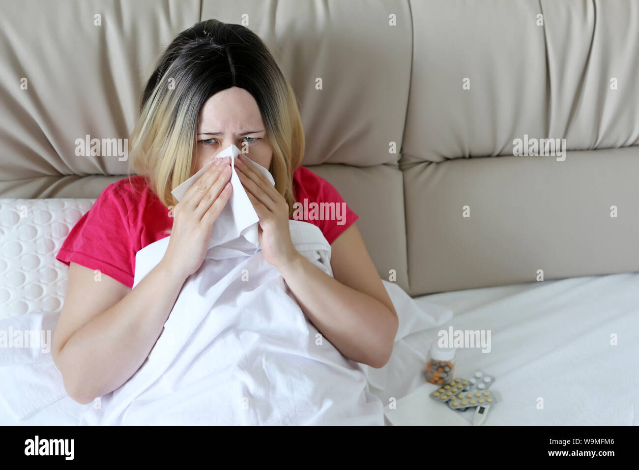 Blonde woman blowing her nose in a handkerchief sitting in a bed. Unhappy girl suffering from runny nose, concept of sick at home, flu, fever Stock Photo