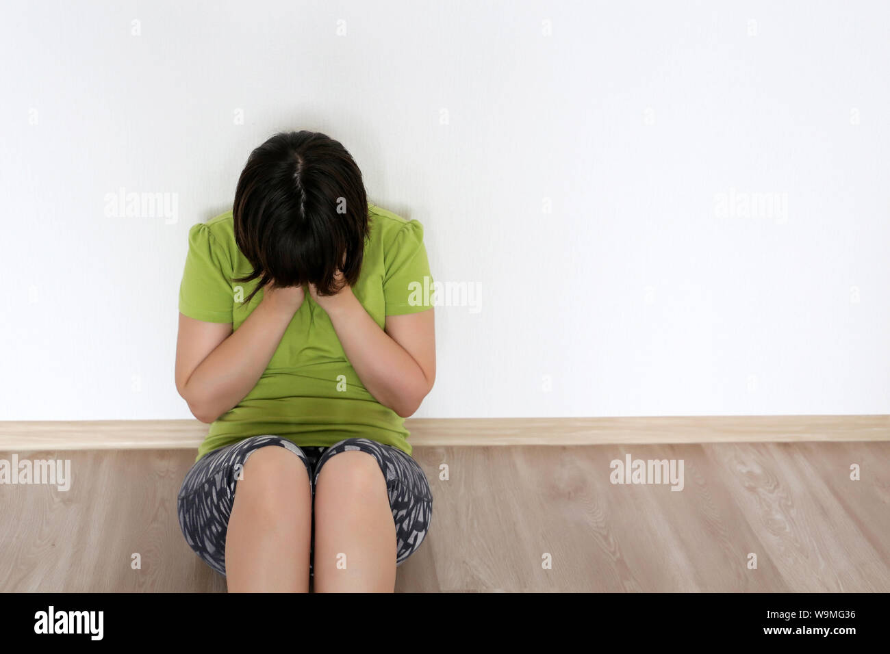 Upset girl sitting on the floor in the room, covering her face with hands. Concept of domestic violence, tragedy, crying woman Stock Photo