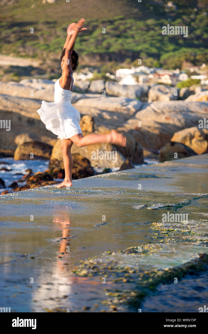 African woman jumping in the air in a summer dress at the beach on a seawall, selected focus on only her face, Camps Bay, Cape Town, South Africa Stock Photo