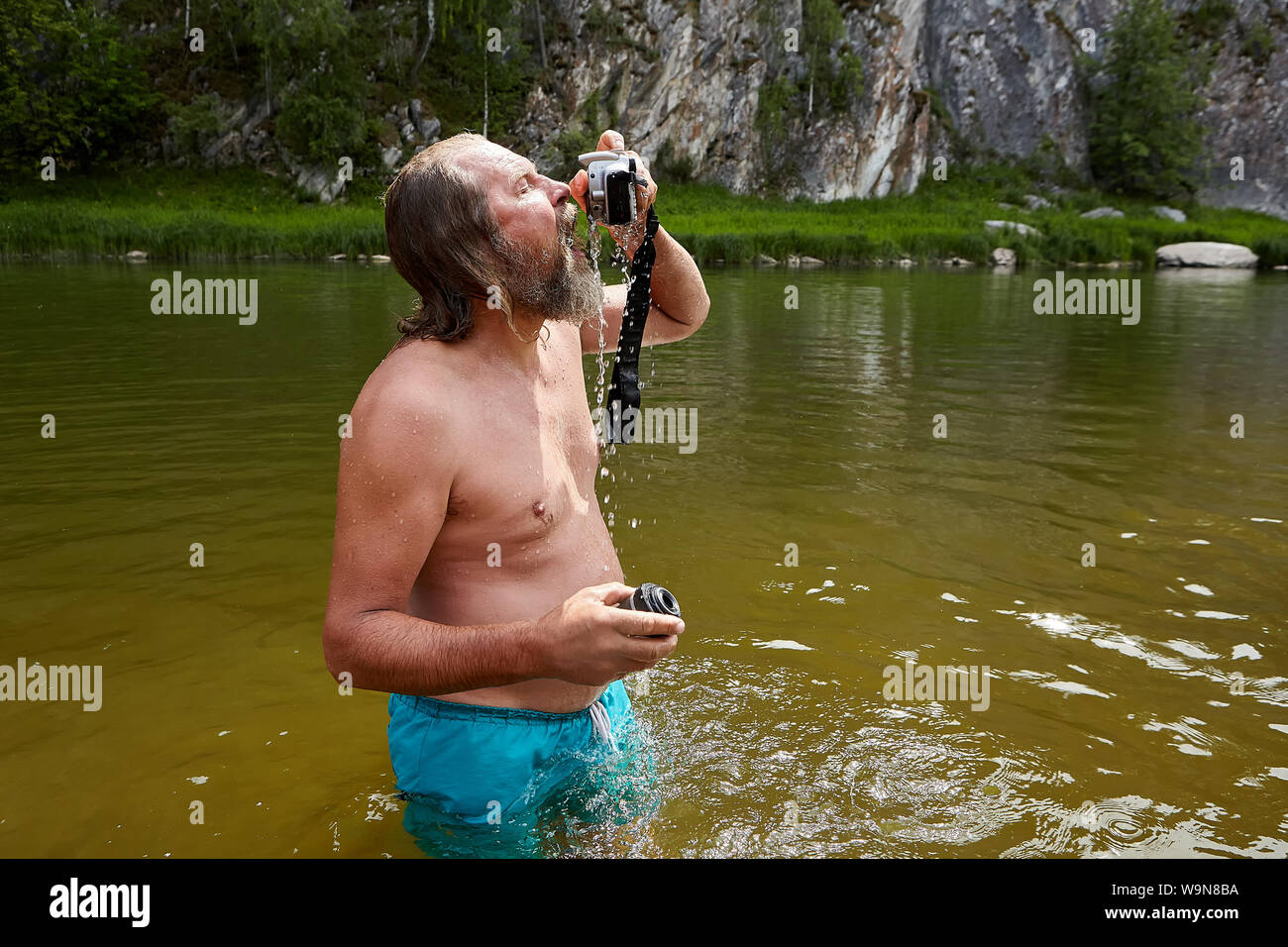 Wet mature bearded man is standing in river with digital camera in his hands. Stock Photo