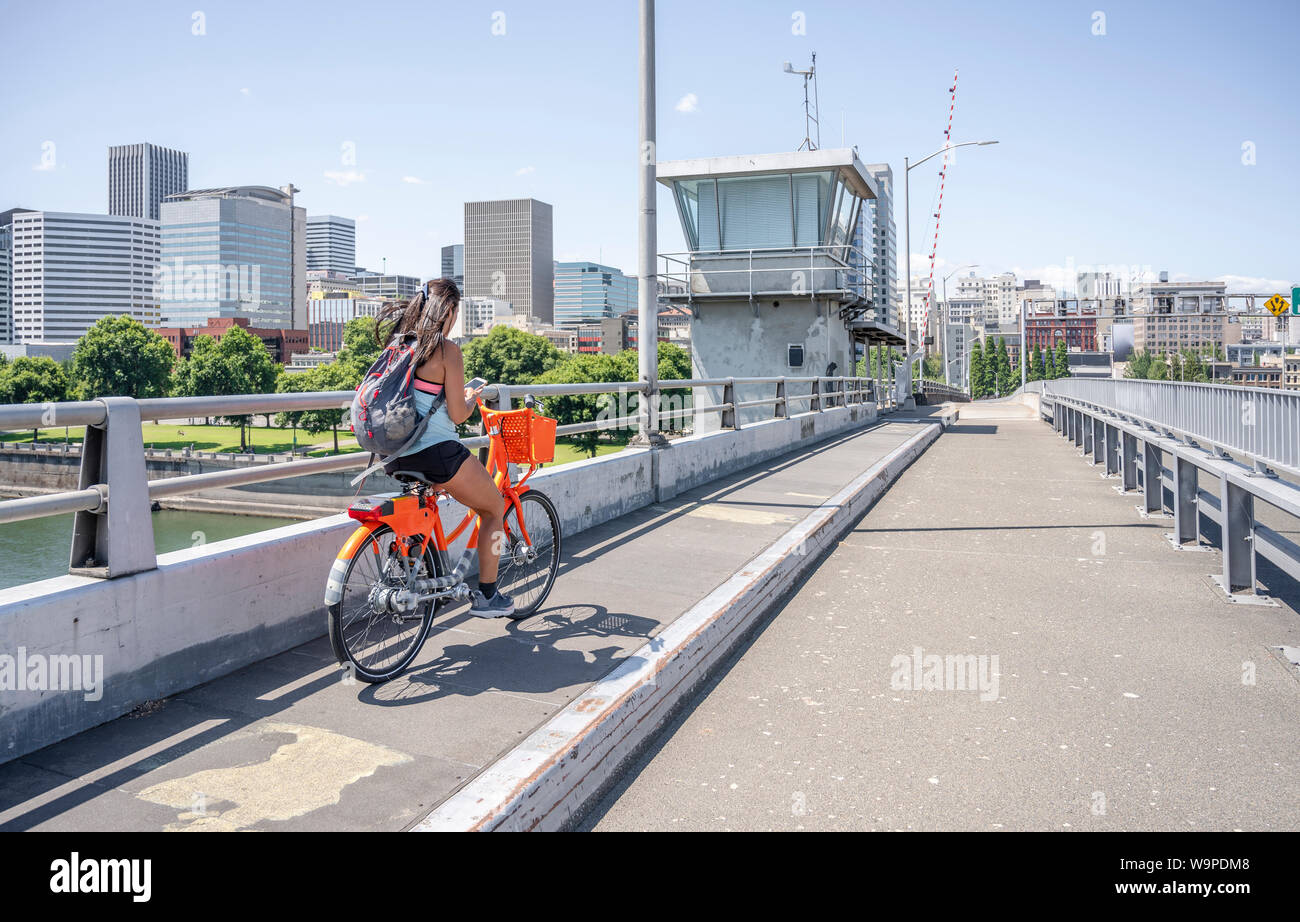 A young girl with her hair and backpack rides an rented orange bike over the bridge and simultaneously flows on the phone, representing the lifestyle Stock Photo
