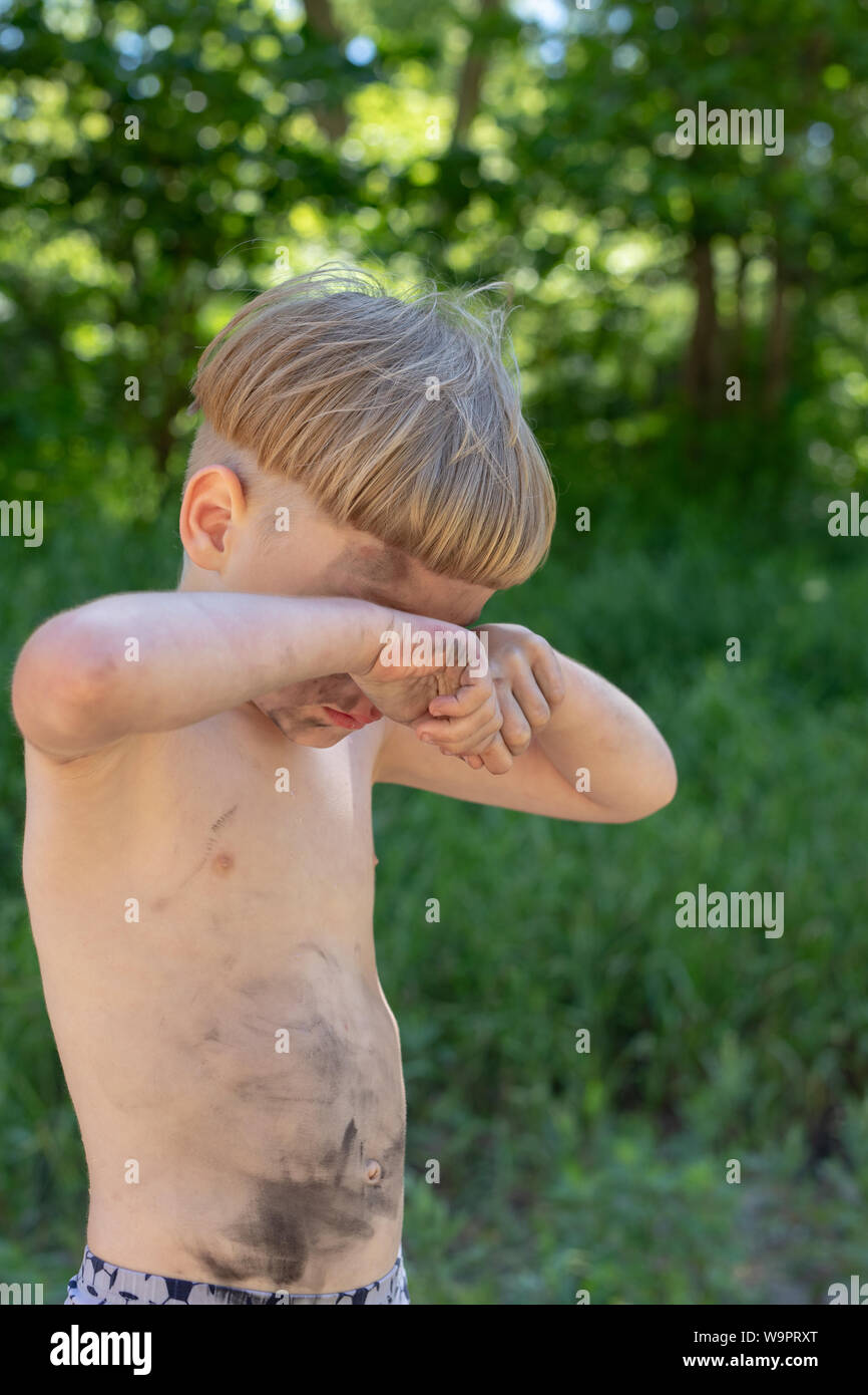 Upset, sad and crying caucasian boy 5 years old with blond hair, stained or dirty. Rubs his eyes. Outside Stock Photo