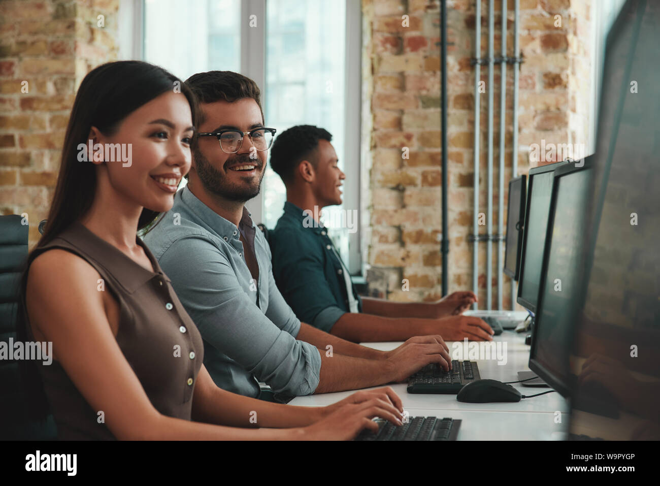 Happy to work together. Group of cheerful young employees working on computers and smiling while sitting in modern open space. Job concept. Business people. Workplace Stock Photo
