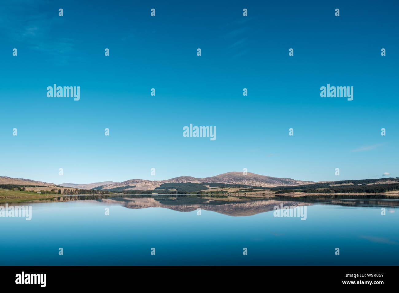 Clatteringshaws Loch in the Galloway Forest in Scotland with the blue skies and hills perfectly reflected in the mirror calm surface of the loch Stock Photo