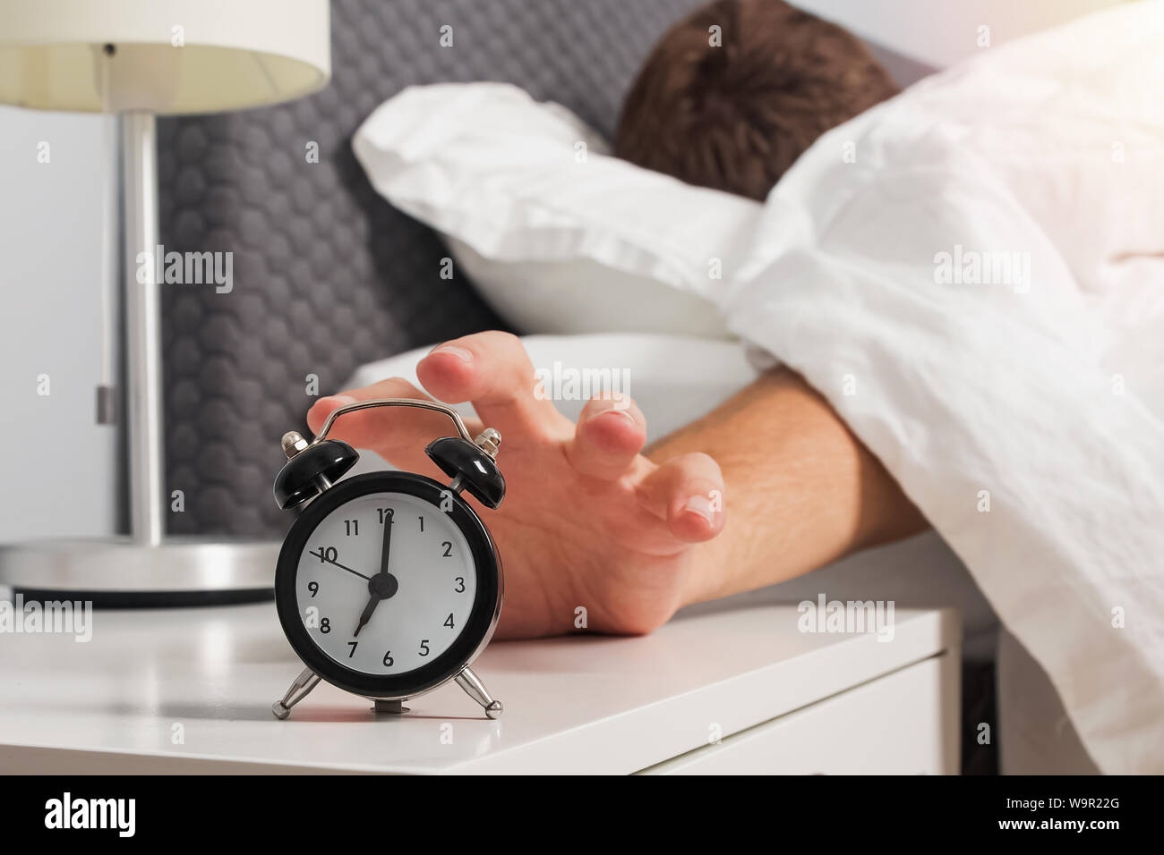 Man's hand reaching out alarm clock on the nightstand early in the morning. Morning routine concept Stock Photo