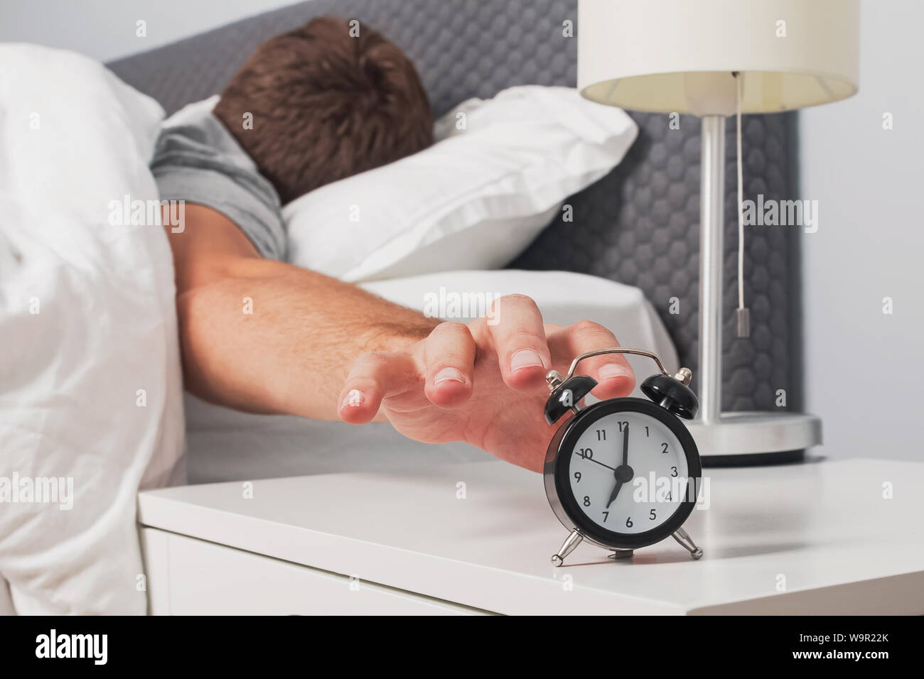 Man's hand reaching out alarm clock on the nightstand early in the morning. Morning routine. Stock Photo