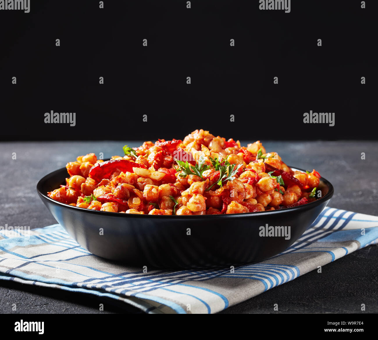 Garbanzos fritos, hot Chickpea stew with sliced chorizo, ham, tomatoes and spices in a black bowl on a concrete table with the black background behind Stock Photo