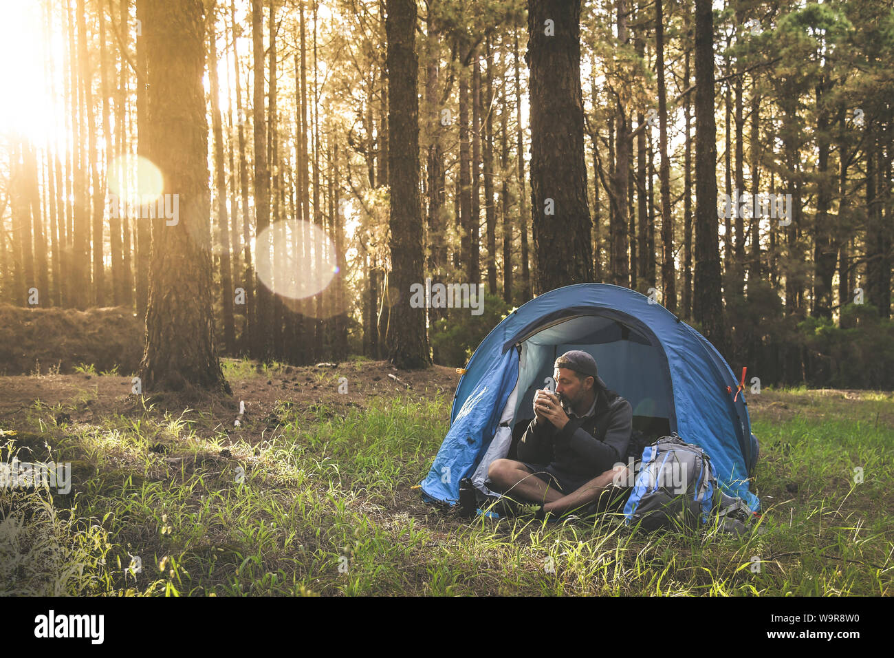 Beautiful young male drinking a cup of coffee in camping tent. Tourist boy enjoying outdoor life in mountain On background trees and setting sun Touri Stock Photo