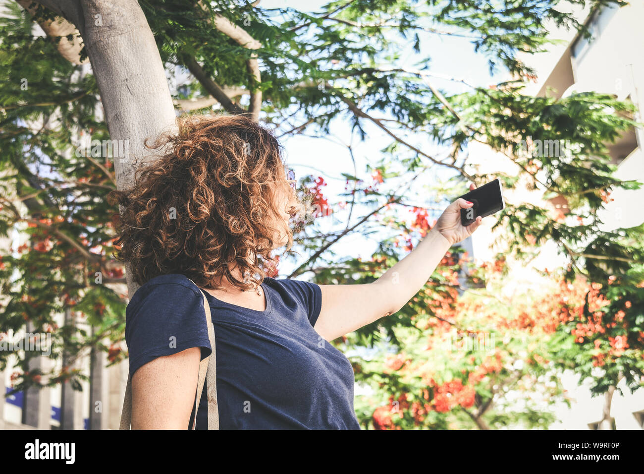 Young woman taking selfie with smartphone outside in the city. Trendy girl playing making new trend video story with cell phone under flowering trees. Stock Photo