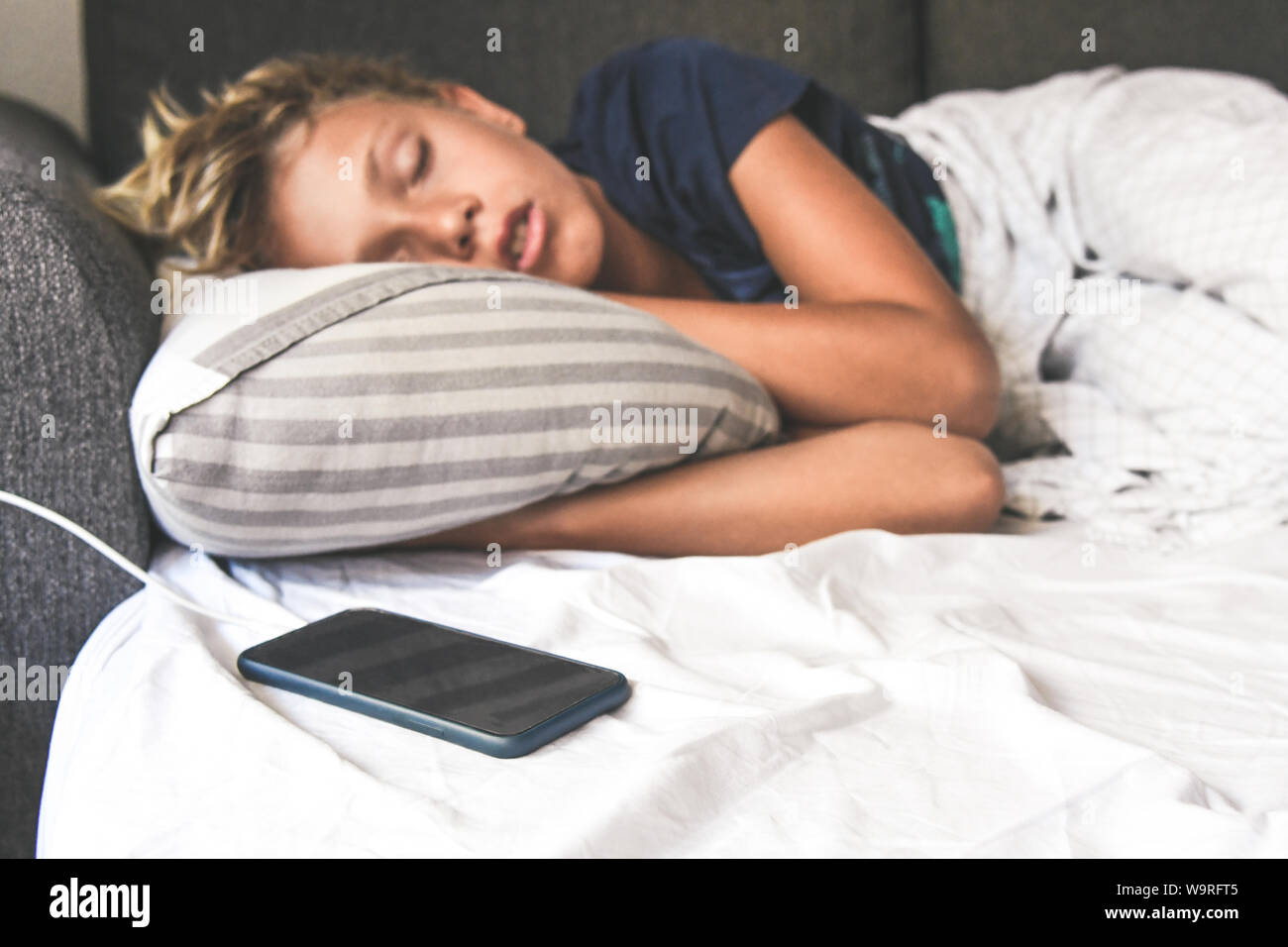 Young boy sleeping with hands under the pillow near the cellphone. Beautiful child sleeps alone in the bed dreaming about online games. Technology add Stock Photo