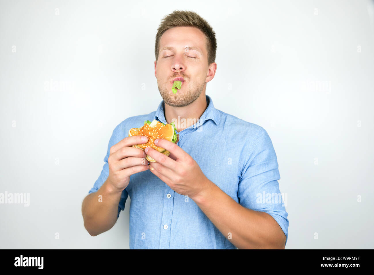 young funny man biting cheeseburger from fast food restaurant standing with salad leaf in his mouth on isolated white background. Stock Photo
