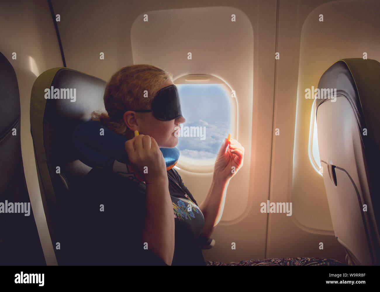 33 year old woman using traveling pillow, sleeping mask and ear plugs in plane cabin on passenger seat, fly with airplane, comfortable cozy traveling Stock Photo