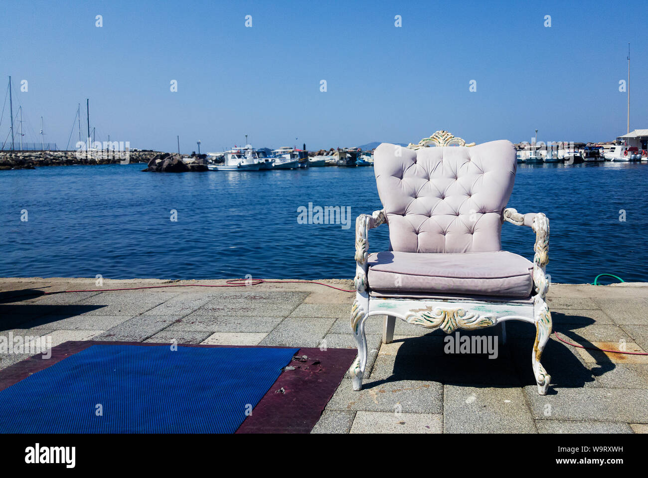 An armchair with baroque decorations standing on cement in the foreground. A fisher port, boats and the Mediterranean sea are seen at the background, Stock Photo