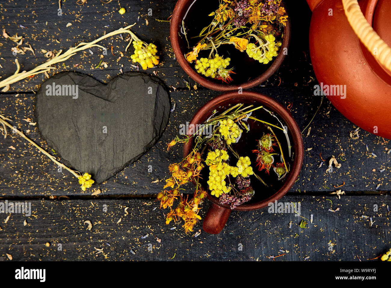Two Cup of herbal tea tutsan, sagebrush, oregano, helichrysum, lavender near brown teapot on dark wooden background. Herbal tea.Flat lay. Hear. Love. Stock Photo