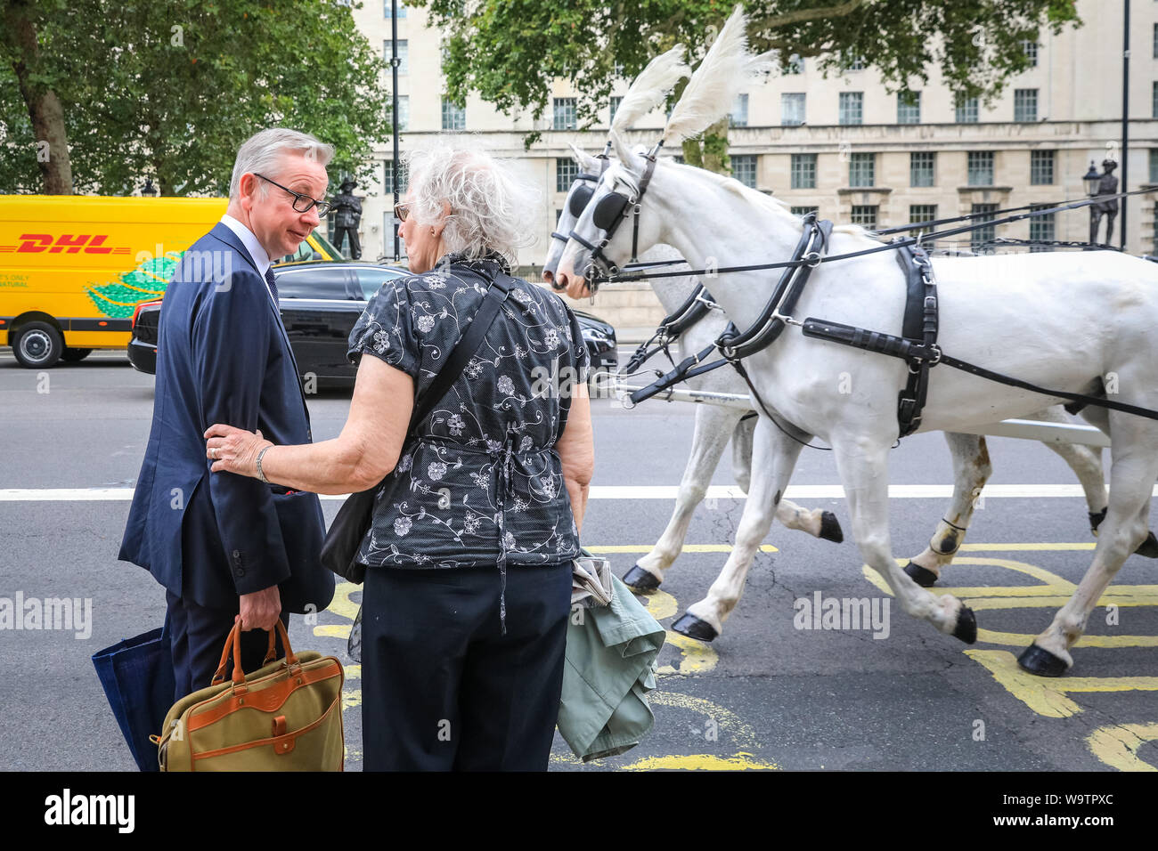 London, UK. 15th Aug, 2019.  Michael Gove, Chancellor of the Duchy of Lancaster with responsibility for no-deal Brexit preparations, as well as overseeing constitutional affairs, maintaining the integrity of the Union and having oversight over all Cabinet Office policy, exits the Cabinet Office in Whitehall and chats to a woman as a horse-drawn carriage goes past them. Credit: Imageplotter/Alamy Live News Stock Photo