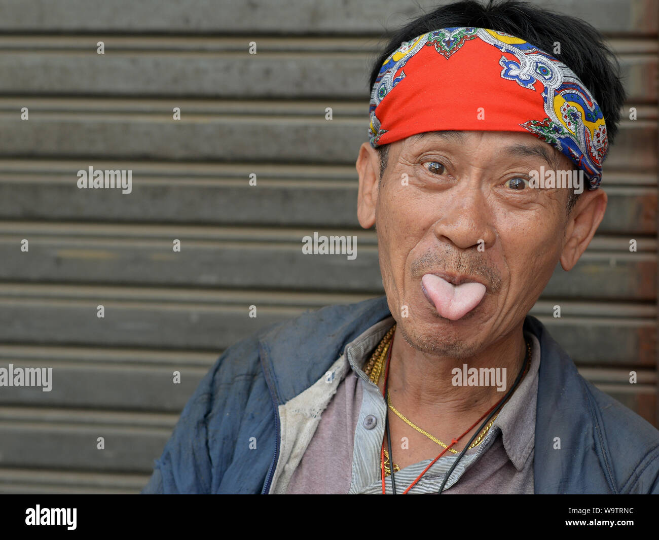 Funny middle-aged Thai man with a colourful bandana sticks out his tongue and mocks the photographer. Stock Photo
