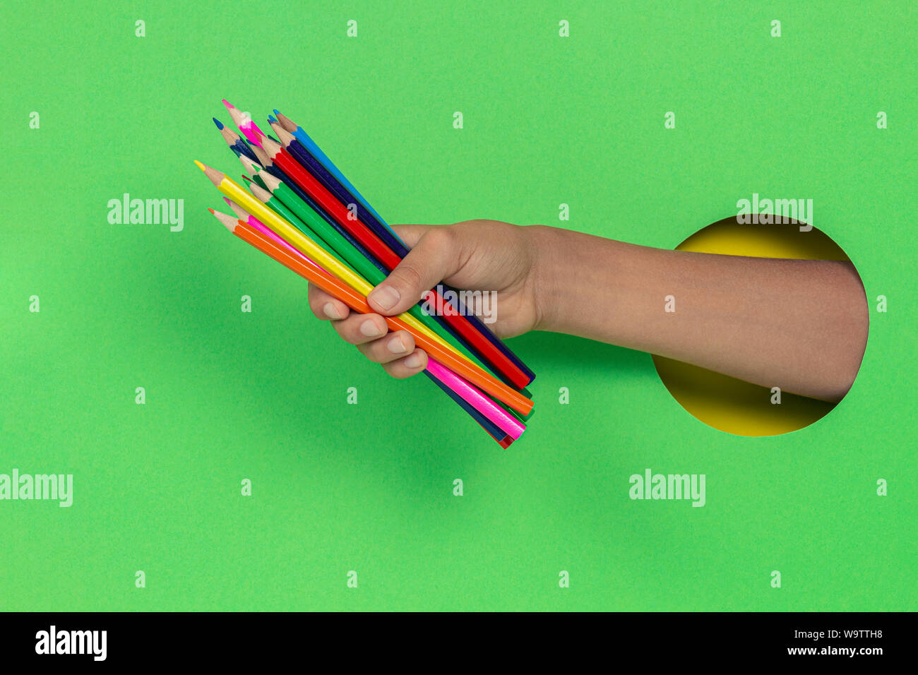 Kid holding colored pencils in hand through hole on light green background Stock Photo