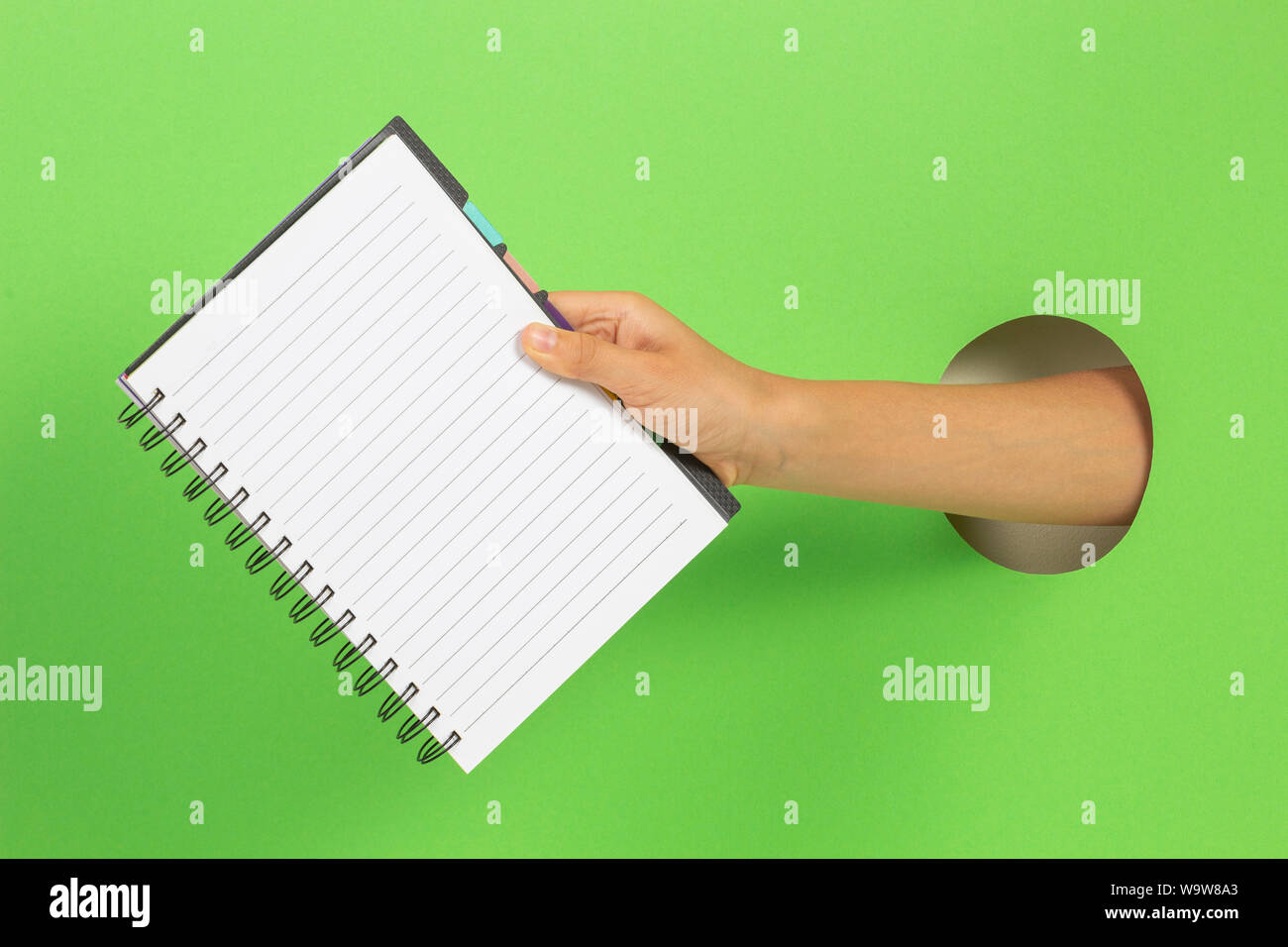 Kid holding open spiral notebook in hand through hole on light green background Stock Photo