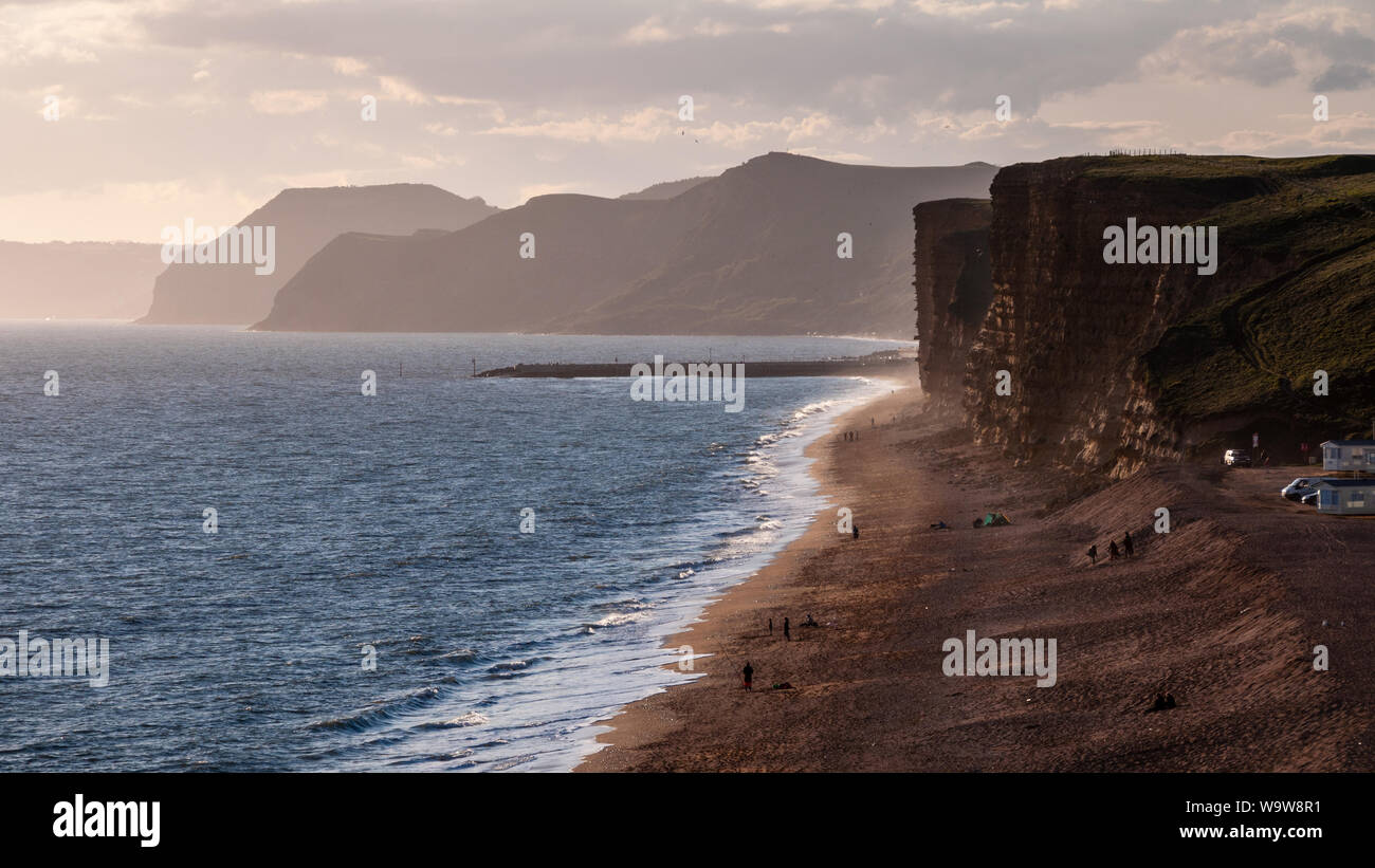 People walking and fishing on Hive Beach at Burton Bradstock are silhouetted against the evening sun on Dorset's Jurassic Coast. Stock Photo