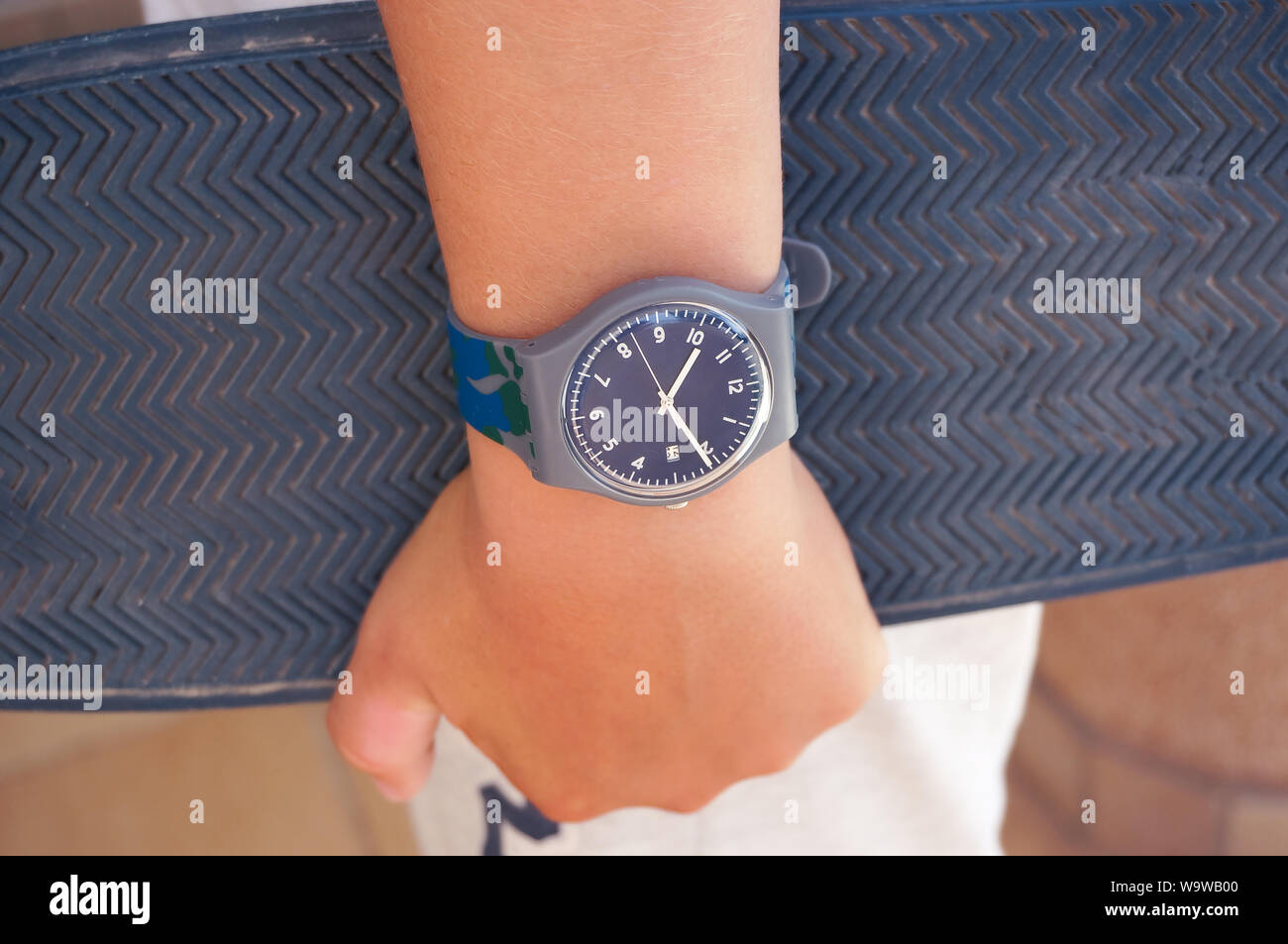 Alicante, Spain - August, 2019: Grey rubber Swatch watch on teenager's wrist holding skate board Stock Photo
